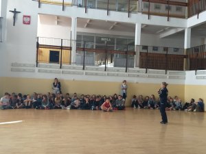 korytarz szkolny policjantka z mikrofonem prowadzi pogadankę, dzieci siedzą na podłodze w okręgu