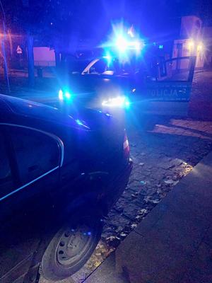 w nocy z lewej strony tył samochodu BMW za nim stojący radiowóz policyjny z włączonymi niebieskimi sygnałami świetlnymi
