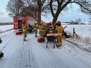 na zaśnieżonej drodze stoi wóz strażacki, z prawej strony przy drzewie samochód osobowy a przy nim strażacy
