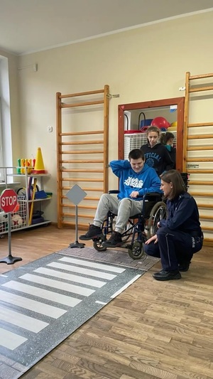na sali chłopiec na wózku inwalidzkim, za nim stojąca dziewczyna, obok wózka klęczy policjantka. Przed chłopcem rozłożona jest mata z przejściem dla pieszych