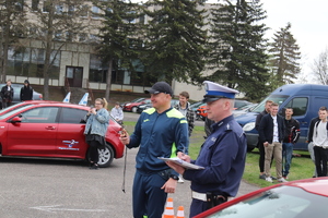 Na boisku stojący policjant obok niego mężczyzna. W tle czerwony samochód osobowy i osoby oglądające