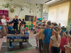 w sali stojące przedszkolaki, z prawej strony dwoje dzieci mają założony element autochodzika radiowóz, przed nimi rozłożona jest jezdnia i obok stoi policjantka