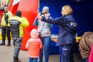 Bezpieczna Majówka w Mikołajkach na fotografii z prawej strony stoi policjantka a obok z lewej strony jest kobieta, która przymierza alkogogle, przy niej stoi odwrócone tyłem dziecko. Z lewej strony obrazka stoi dwóch mężczyzn