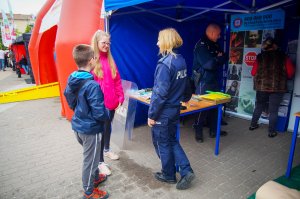 Bezpieczna Majówka w Mikołajkach na fotografii przy policyjnym stanowisku umiejscowionym pod niebieskim namiotem stoi policjantka i rozmawia z dwójką dzieci