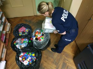 Na zdjęciu jest policjantka, która z kartonu przesypuje zebrane plastikowe nakrętki do worka z nakrętkami, przed nią stoją cztery worki z nakrętkami