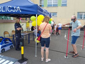 Festyn w Mikołajkach,  stanowisko policji, z lewej strony niebieski namiot policyjny, pod nim stoły a na nic h ulotki, przed czerwone słupku i mężczyzna w alkogoglach pokonuje tor przeszkód.