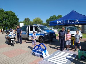 na placu szkolnym z prawej strony policyjny namiot i autochodzik i policjantka z dziećmi ucząca przechodzić przez przejście dla pieszych, z lewej strony policyjny bus i motocykl na którym siedzi chłopiec, a obok stoi policjant