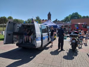 na placu szkolnym z prawej strony policyjny motocykl, obok policjant, z lewej strony radiowóz bus a w środku dzieci