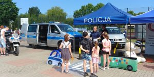 na placu szkolnym z prawej strony policyjny namiot i autochodzik i policjantka z dziećmi ucząca przechodzić przez przejście dla pieszych, z lewej strony policyjny bus i motocykl