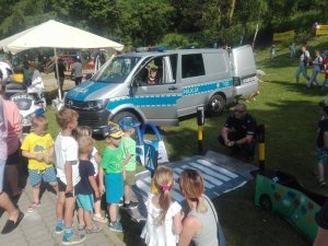 na palcu przy przedszkolu na trawie z prawej strony kuca policjant przy zestawie autochodzik a z drugiej strony stoją dzieci, w tle stoi radiowóz bus