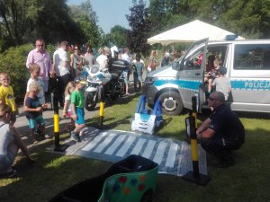 na palcu przy przedszkolu na trawie z prawej strony kuca policjant przy zestawie autochodzik a z drugiej strony stoją dzieci przed przejściem dla pieszych i jedno przechodzi przez nie, w tle stoi radiowóz bus