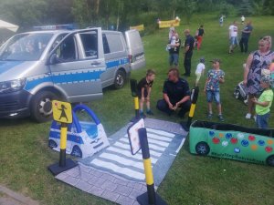 na palcu przy przedszkolu na trawie z prawej strony kuca policjant przy zestawie autochodzik obok niego  dziecko,  w tle stoi radiowóz bus