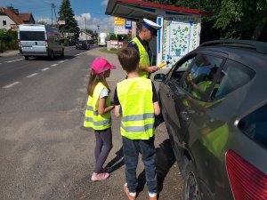 ulica w Marcinkowie z prawej strony auto zatrzymane przez policjanta do kontroli drogowej. policjant trzyma w ręku alcoblow a obok stoi 2 dzieci i trzymają ulotki