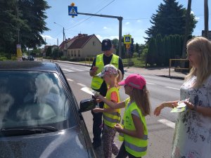 ulica w Marcinkowie z lewej strony stoi auto zatrzymane przez policjanta do kontroli drogowej przy nim stoi policjant 2 dzieci i nauczycielka, dzieci podają ulotki dla kierowcy