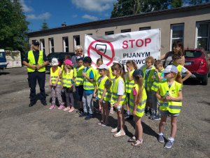 zdjęcie grupowe uczestników działań, dzieci w żółtych odblaskowych kamizelkach stoją z policjantem i nauczycielami za nimi stoi baner z napisem stop pijanym kierowcom