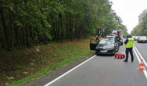 trasa k 16 jezdnia, z lewej strony ściana lasu na lewym pasie drogi  stoi auto marki toyota z uszkodzeniami przodu i szyby, przed autem stoi policjant,  przy aucie stoją strażacy i z tyłu ambulans, z prawej strony na pasie stoją inne auta