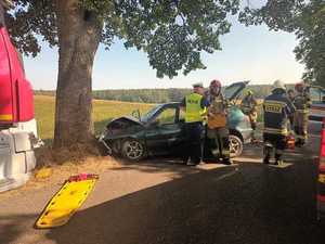 samochód z rozbitym przodem stojący przy drzewie, stojący przy nim policjant i strażacy