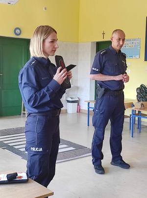 Z lewej strony stoi policjantka trzymająca w rękach element autochodzika- sygnalizacja świetlna a obok niej stoi policjant. Osoby znajdują się w szkolnej sali, za nimi na podłodze leży rozłożona plansza z przejściem dla pieszych