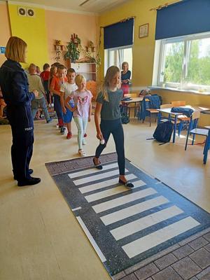 Z lewej strony stoi policjantka a na podłodze leży rozłożona plansza z przejściem dla pieszych na której przechodzą dzieci. Osoby znajdują się w szkolnej sali.