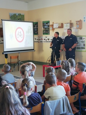 dwóch policjantów stojących przy tablicy na której wyświetlany jest znak drogowy zakaz wjazdu rowerom i siedzące w sali dzieci