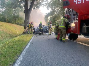 z lewej strony widoczne drzewo przy drodze, przy nim leży rozbite auto, przy aucie stoją strażacy, z prawej strony zdjęcia widać wóz strażacki i strażak