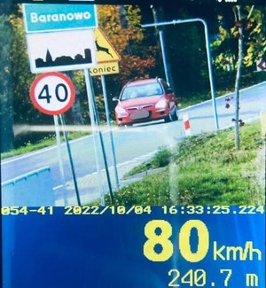 z lewej strony znajduje się znak drogowy z ograniczeniem prędkości do 40km/h, znak drogowy teren zabudowany oraz nazwa miejscowości Baranowo. Na drodze widać jadący czerwony samochód. Pod spodem żółty napis 80 km/h