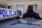 kogut na dachu radiowozu policyjnego, policjant stojący przodem do budynku komendy a tyłem do zdjęcia