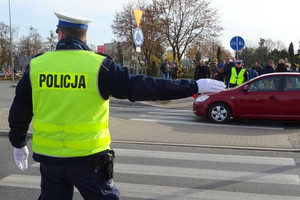 stojący na skrzyżowaniu policjanci ruchu drogowego ubrani w żółte kamizelki, kierujący ruchem pojazdów, nadjeżdżający z prawej strony samochód, w tle widoczny cmentarz