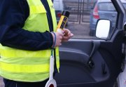z lewej strony stoi policjant w żółtej kamizelce w ręce trzyma alkomat i "lizak policyjny", stoi przed otwartymi drzwiami samochodu