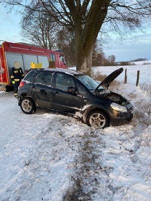 na zaśnieżonej drodze częściowo w rowie przy drzewie stoi samochód osobowy z uszkodzonym przodem, w tle wóz strażacki i strażacy