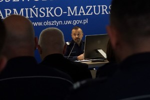 mężczyźni w mundurach siedzący tyłem w tle Komendant Wojewódzki Policji w Olsztynie przemawiający do mikrofonu