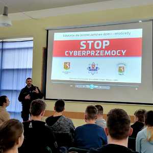 policjant omawiający wyświetlaną prezentację z napisem stop cyberprzemocy przed nim siedząca młodzież