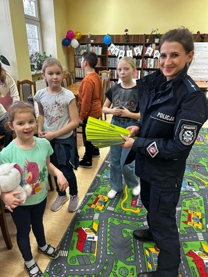 w bibliotece policjantka stojąca obok dzieci trzymająca w ręce opaski odblaskowe