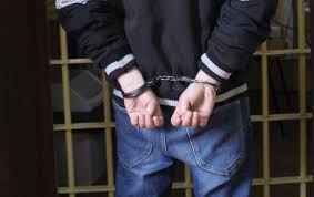 stojąca tyłem osoba na tle krat z założonymi kajdankami na ręce trzymane z tyłu