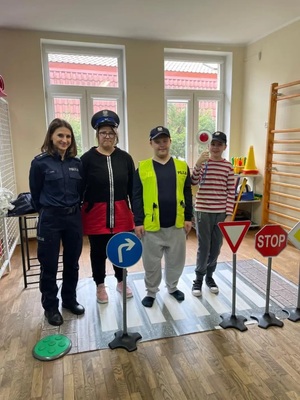 na sali stojąca z lewej strony policjantka obok troje dzieci ubranych w elementy ubioru policyjnego, przed nimi stoją znaki drogowe