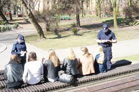 sześć osób siedzących w parku na ławce, przed nimi stoi dwóch policjantów i ich legitymują