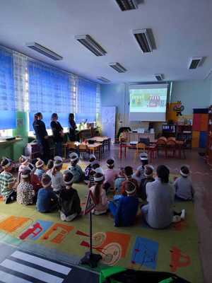 w sali siedzące na dywanie dzieci oglądające wyświetlany na ekranie filmik, po lewej stronie stoją policjantki i opiekunka