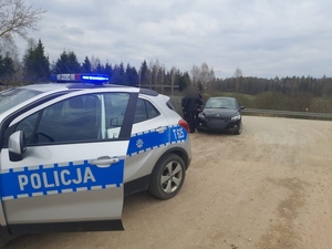 radiowóz policyjny z włączonymi sygnałami świetlnymi a za nim stojący mężczyzna, kobieta i policjant obok samochodu koloru czarnego