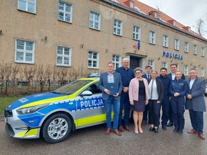Przed budynkiem Komendy Powiatowej Policji w Mrągowie stoi zaparkowany oznakowany radiowóz, a przy nim stoją samorządowcy, Komendant Powiatowy Policji w Mrągowie i policjantka