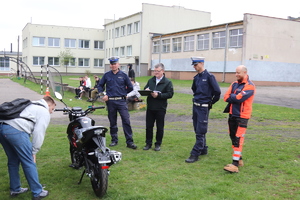 na terenie zewnętrznym stoi dwóch policjantów, ratownik medyczny i mężczyzna. Przed nimi stoi motorower, przed którym pochyla się chłopak