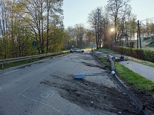 leżąca częściowo na pasie zieleni a częściowo na jezdni przewrócona latarnia, w oddali widać stojący w poprzek drogi rozbity samochód osobowy