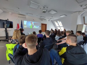siedząca na sali odpraw młodzież oglądająca wyświetlany film o służbie w Policji