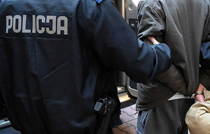 policjant prowadzący zatrzymanego, który ma założone kajdanki na ręce trzymane z tyłu