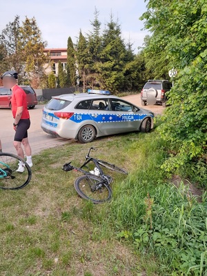 w okolicach ogródków działkowych, częściowo na trawie stoi rower, obok niego stoi mężczyzna ubrany w strój rowerowy, obok na trawie leży drugi rower, przy nim stoi radiowóz policyjny