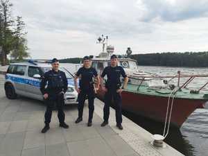 na promenadzie stoi dwóch policjantów i policjantka w tle widać radiowóz policyjny a z prawej strony na jeziorze łódź