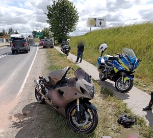 stojący na poboczu drogi uszkodzony motocykl, obok stoja motocykle policyjne, w oddali widać szary samochód osobowy