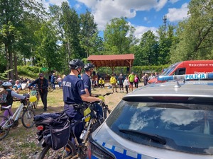 z prawej strony radiowóz policyjny, obok stoją dwaj policjanci z rowerami policyjnymi, w tle piknik rodzinny