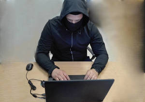 mężczyzna ubrany na czarno z kapturem i maską na twarzy siedzący przed laptopem
