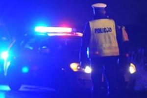 nocą policjant ruchu drogowego w odblaskowej kamizelce a w tle radiowóz policyjny z włączonymi sygnałami świetlno-dźwiękowymi