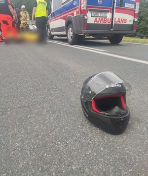 na drodze leżący kask w tle karetka pogotowia stojąca na drodze, obok niej policjant i ratownicy medyczni